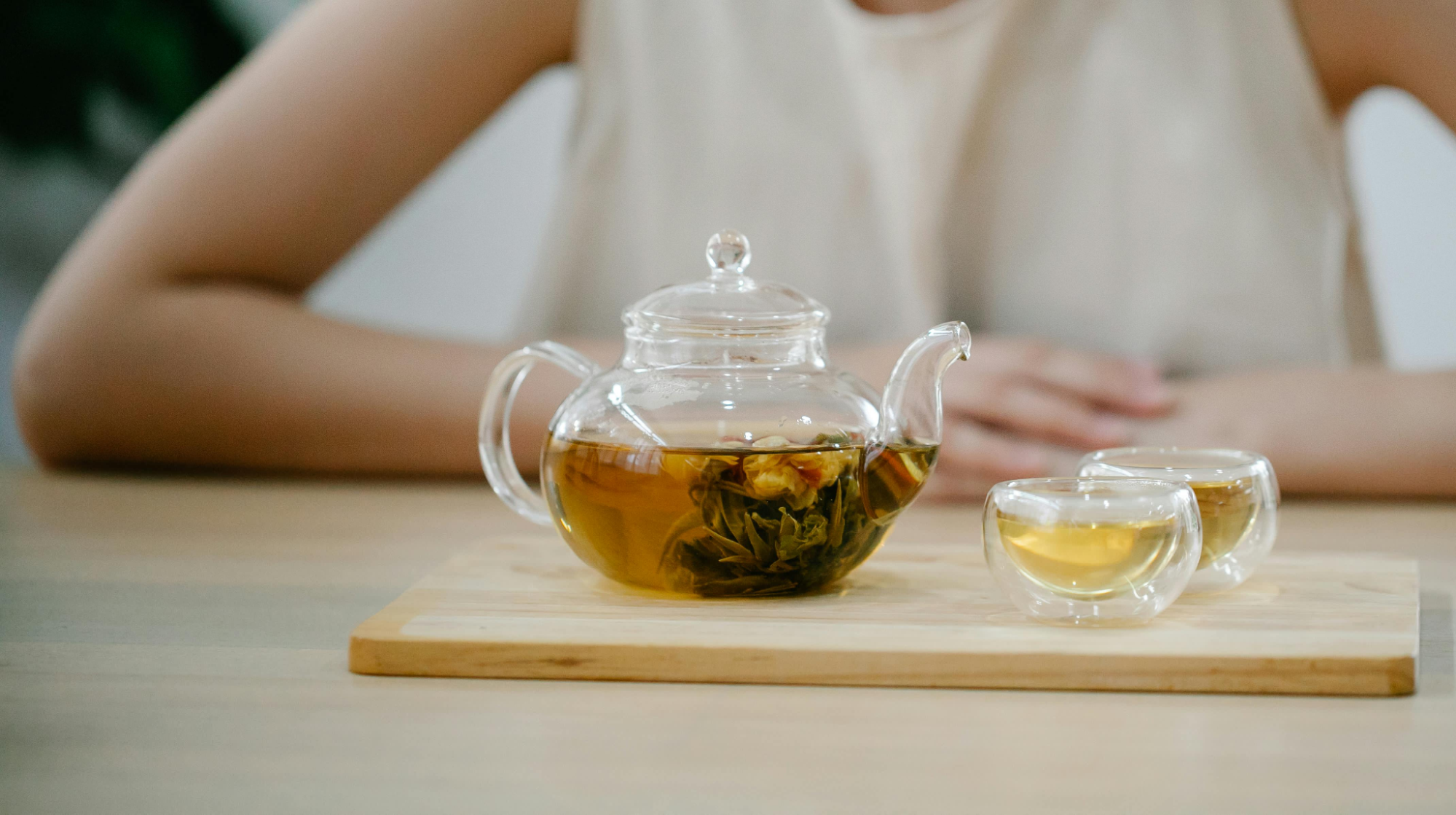 A clear glass teapot with a blossoming green tea flower inside, alongside two small glass tea cups