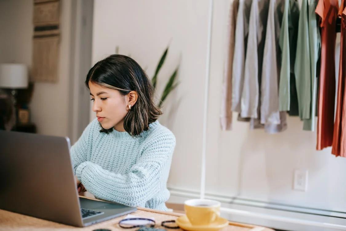 a woman wearing a sweater is working on a laptop at a wooden table with a yellow cup